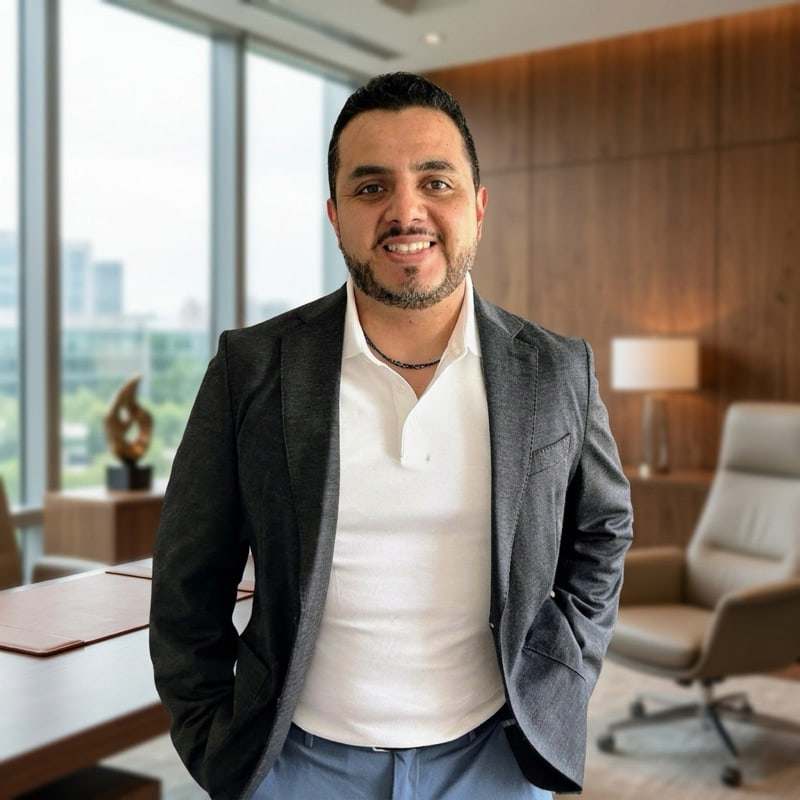 Mauro Flores, standing in a meeting room in a charcoal jacket and an open-necked white shirt, with a city skyline through the window behind him.